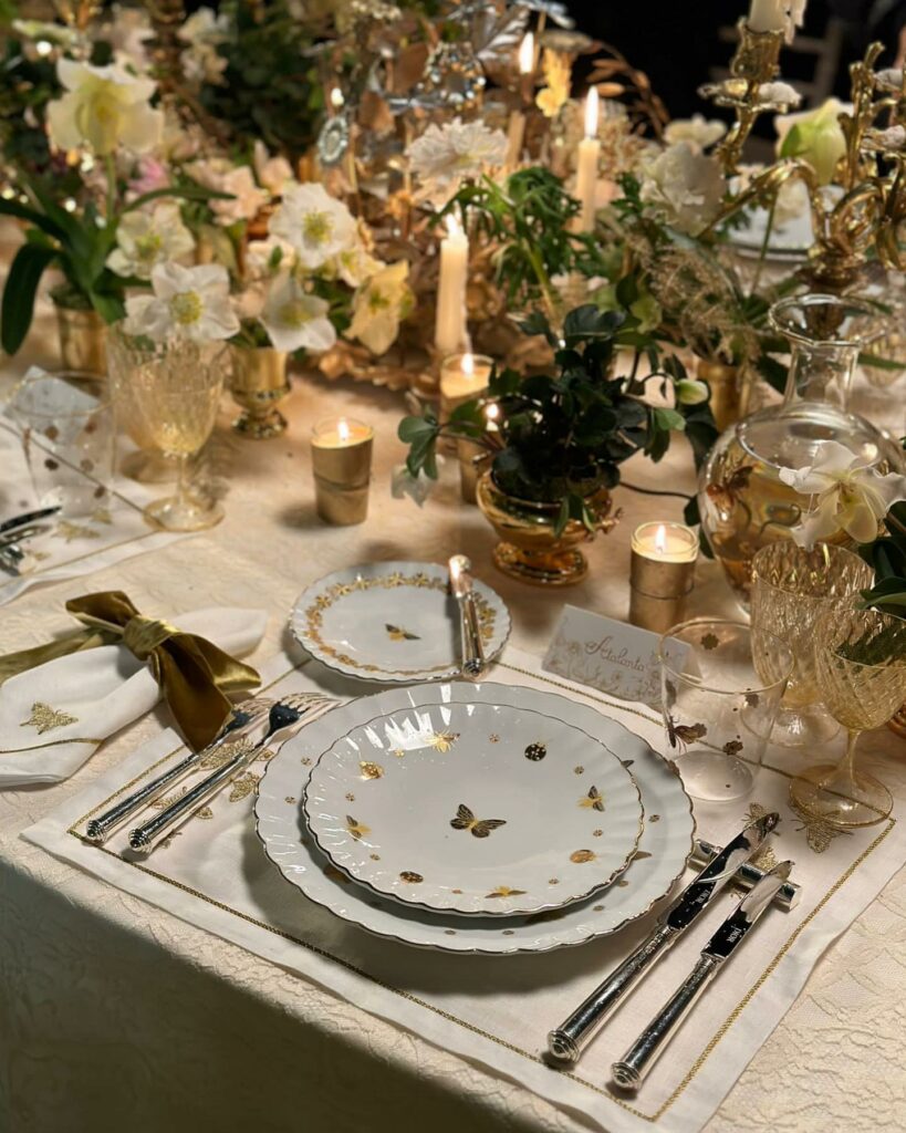 A busy table setting with lots of white, brown, and yellow flowers, a set of white plates, silverware, a white placemat, and candles lit throughout the space.