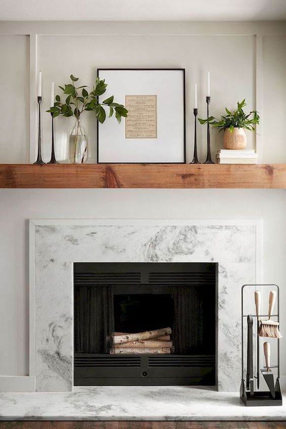A marble fireplace sits below a brown wood mantle topped with various size candles and plants.