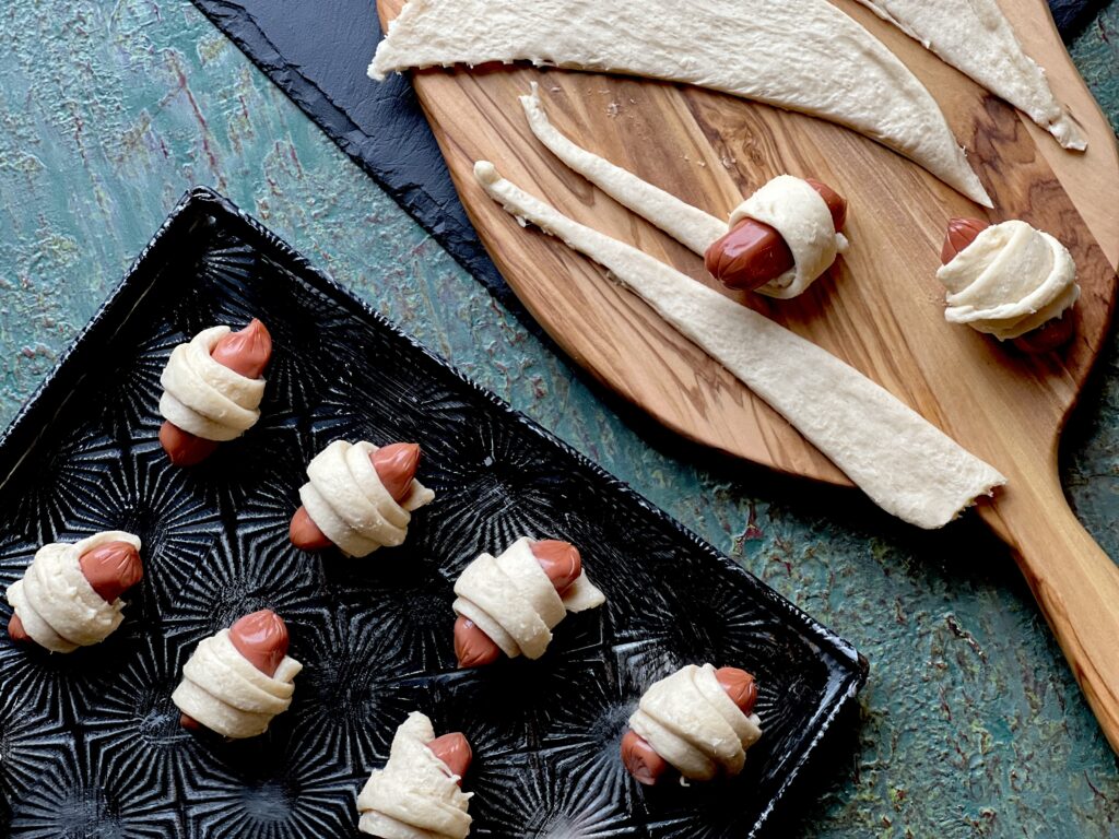 A tray of mini pigs in a blanket ready for the oven, and a wooden board with crescent dough and mini weenies being wrapped.