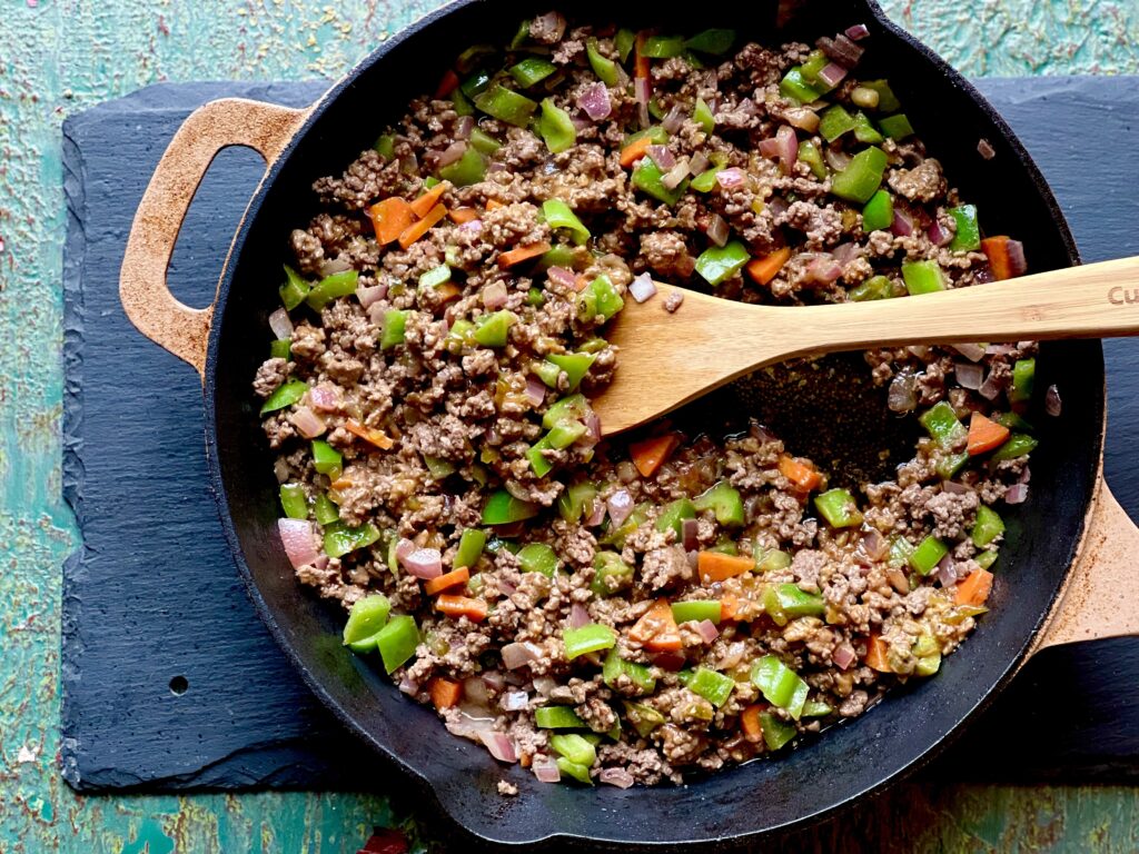 A cast iron skillet with sautéed veggies and ground meat on a piece of slate on a blue/grey surface.