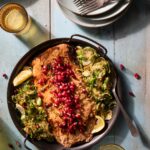 A pan filled with brussel sprouts, salmon, and pomegranate seeds sits on a wooden table surrounded by plates and forks.