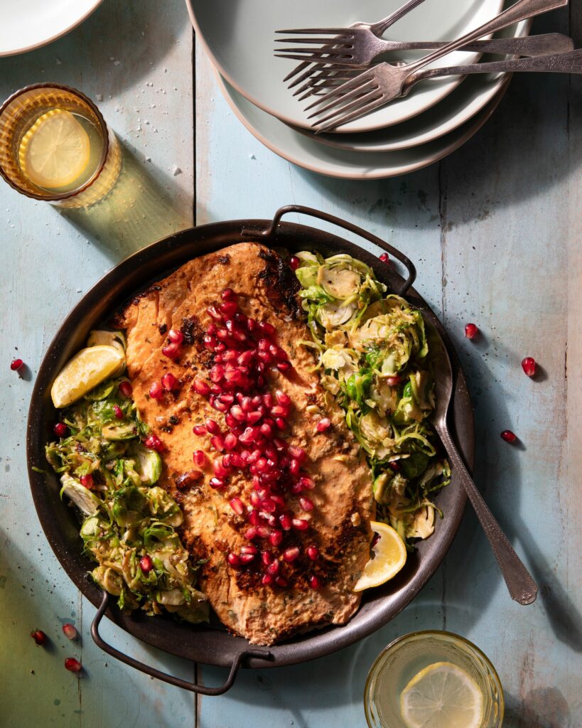 A pan filled with brussel sprouts, salmon, and pomegranate seeds sits on a wooden table surrounded by plates and forks.