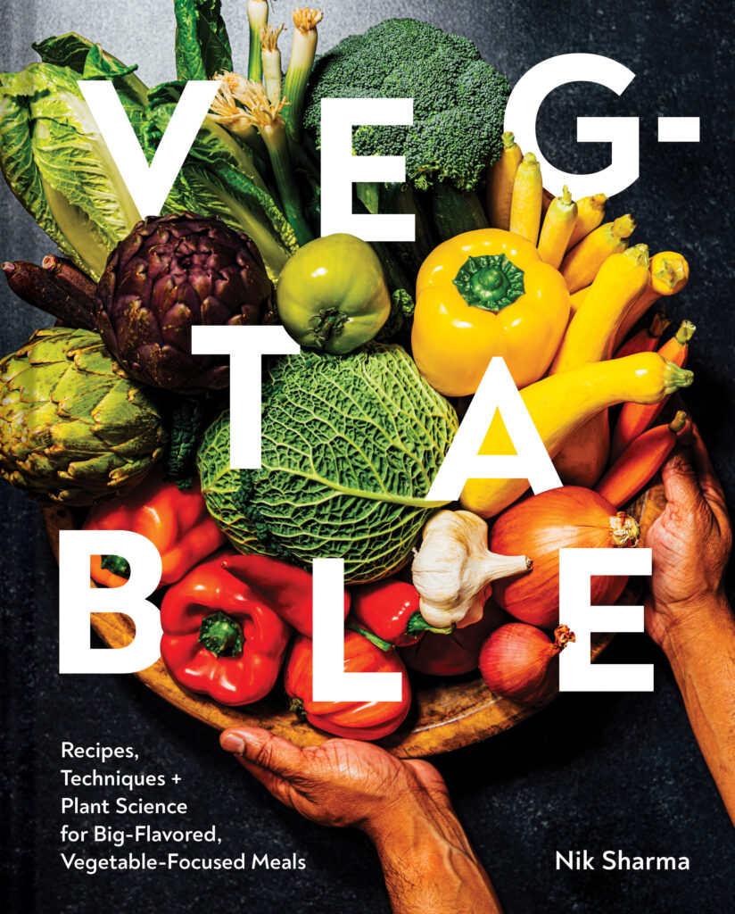 A person holds a basket of various colored vegetables like green cabbage, yellow peppers, onions, and red peppers. The image is placed as the cover of best 2023 cookbook, Veg-Table.