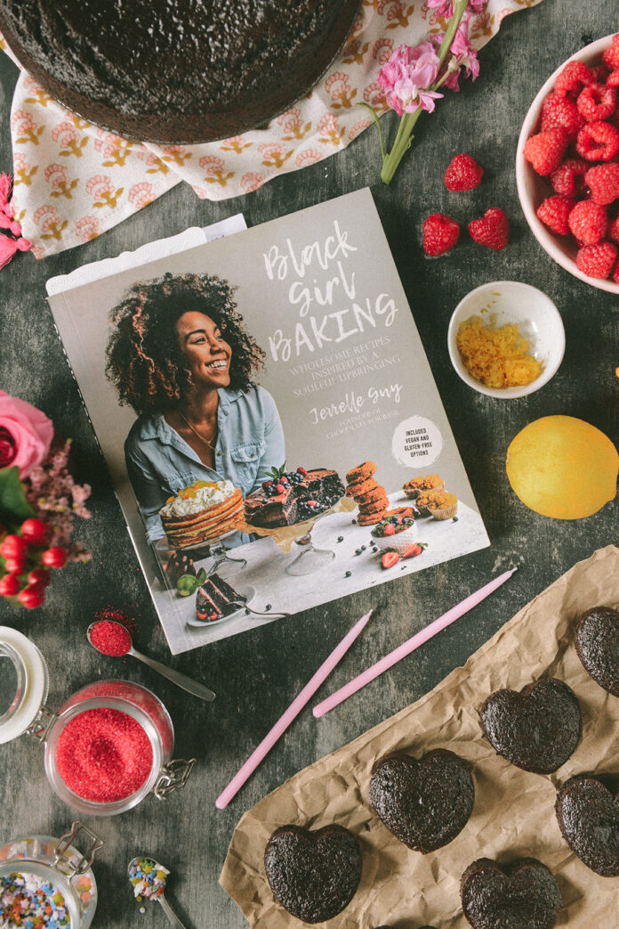 An aerial shot of Black Girl Baking cookbook, various baking ingredients, heart chocolate cupcakes, and raspberries all on a table/