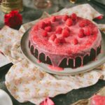 A chocolate cake covered in raspberry glaze and raspberries from Jerrell Guy's cookbook "Black Girl Baking" sits beside a set of cupcakes and other pink decorations.