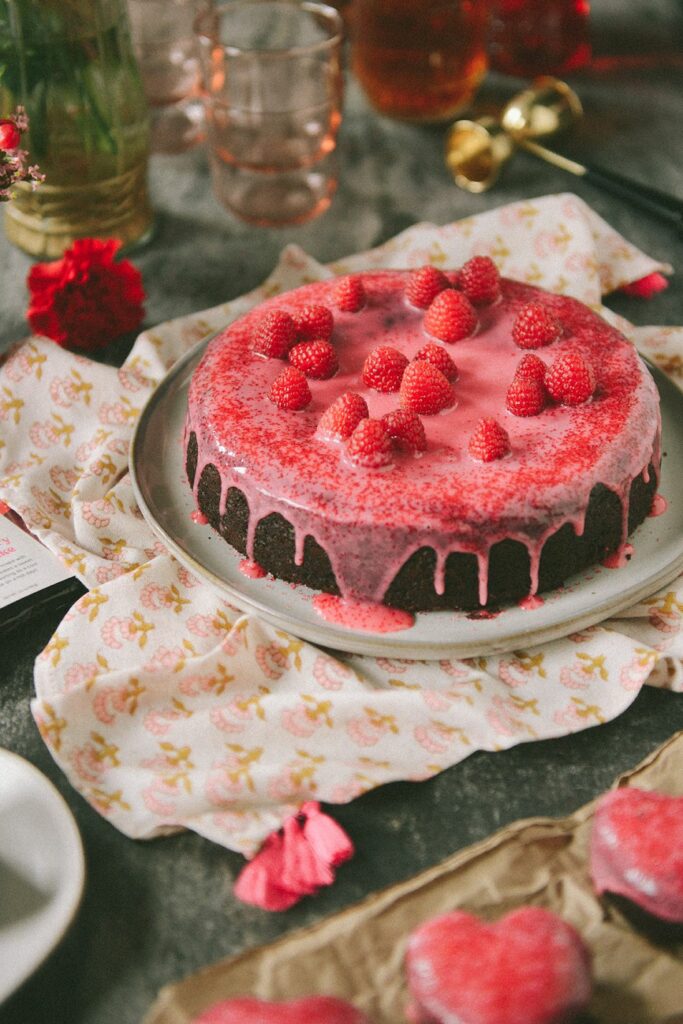 A chocolate cake covered in raspberry glaze and raspberries from Jerrell Guy's cookbook "Black Girl Baking" sits beside a set of cupcakes and other pink decorations.