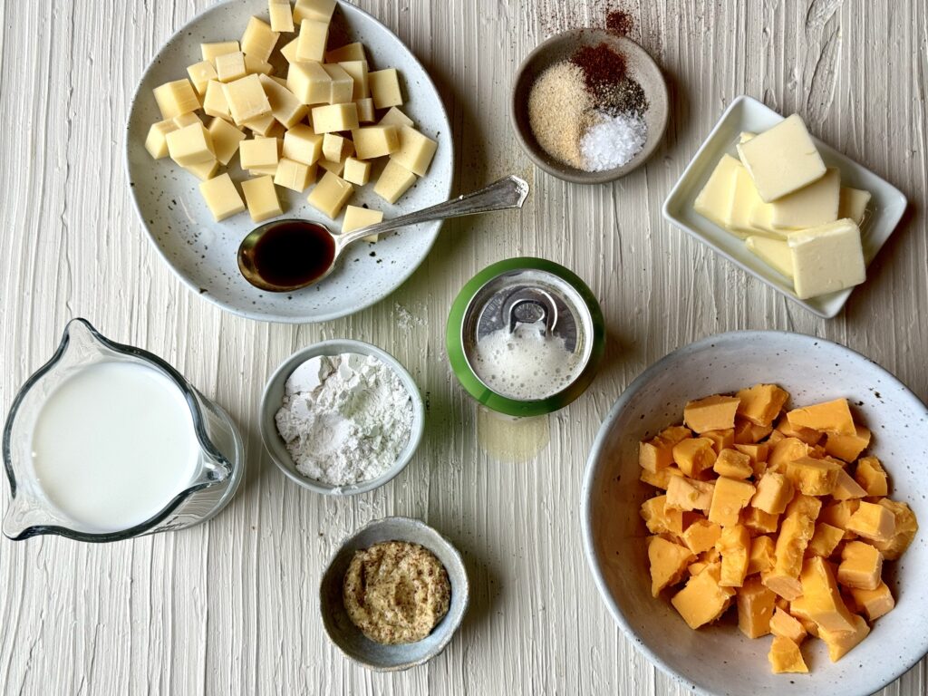 A grouping of ingredients including, milk, beer, cheese, butter, and seasonings in varied sizes of bowls on a textured cream colored surface.