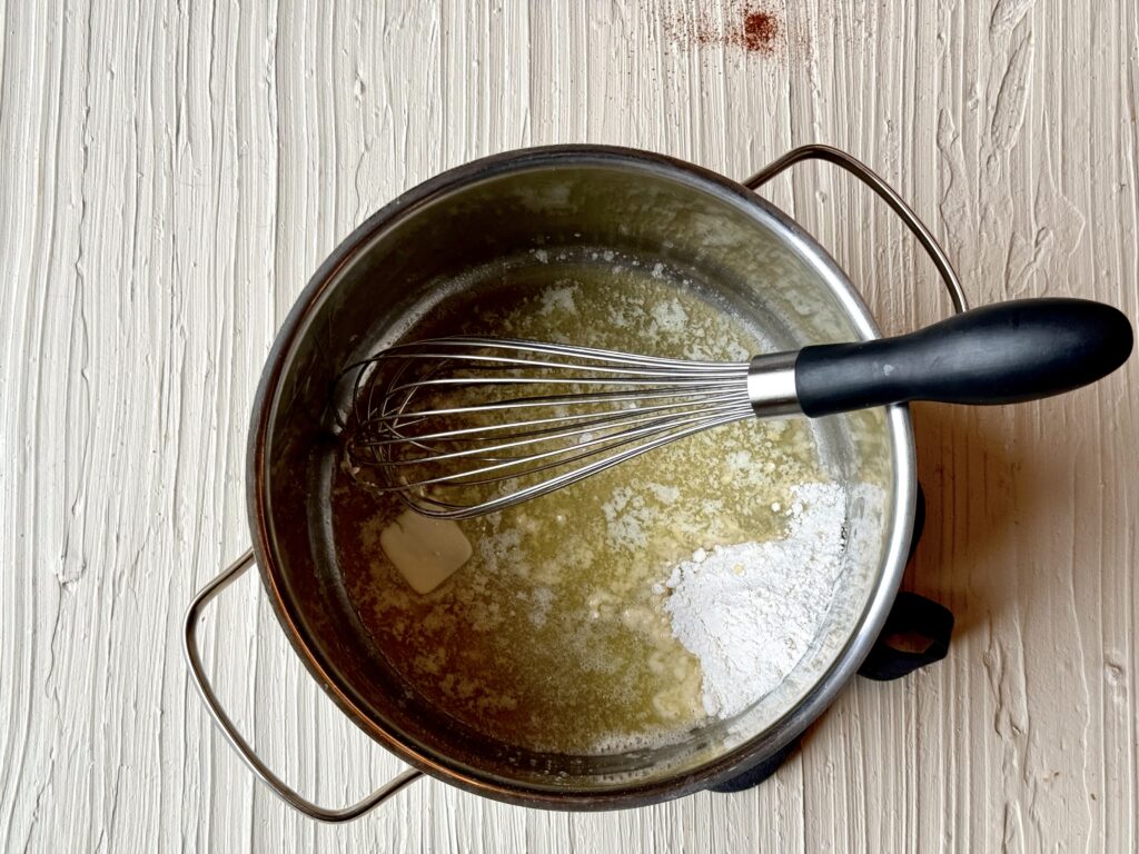 A medium saucepan with melted butter and a whisk on a textured cream colored surface.