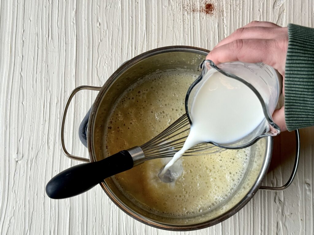 The hand of a women pouring milk into a medium saucepan with with melted butter and flour.