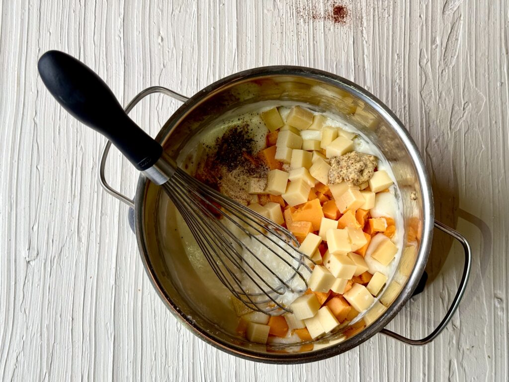 A medium saucepan with cheeses, spices and a whisk on a cream colored texture surface.