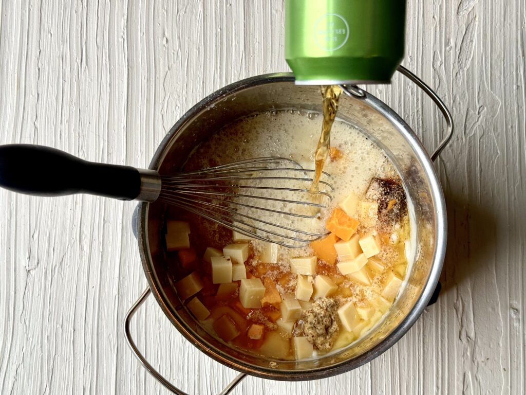 A can of IPA beer being poured into a saucepan with cheese and seasonings on a textured ivory surface.