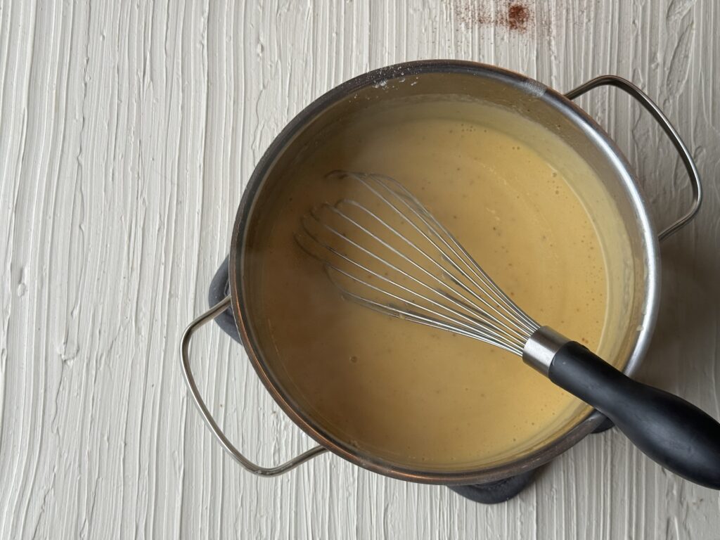 A medium saucepan with a smooth cheese sauce and a whisk on a textured cream surface.