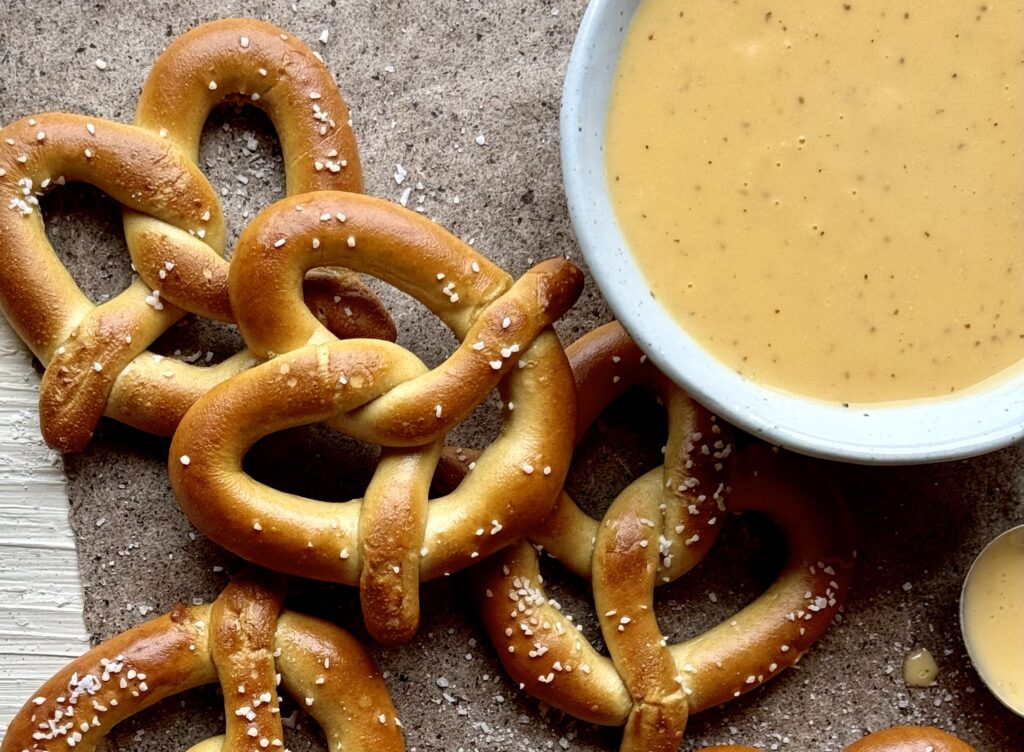 A grouping of soft pretzels next to a white bowl with orange beer cheese on a brown paper surface.