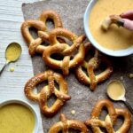 Six soft pretzels on a brown paper with a bowl of orange beer cheese, and a bowl of yellow honey mustard all on a white textured surface.