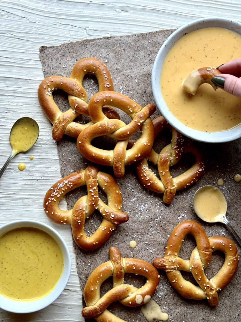 Six soft pretzels on a brown paper with a bowl of orange beer cheese, and a bowl of yellow honey mustard all on a white textured surface.