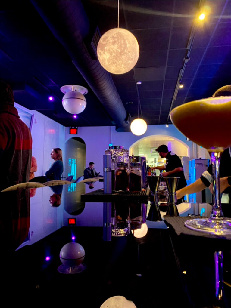 The bar at Space Bar in PIttsburgh has bartenders hard at work on the right side and people enjoying drinks on the left side with a big moon light overhead.