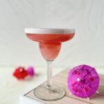 A pink cocktail in a classic margarita glass on a tray with cocktail umbrellas in hot pink and red.