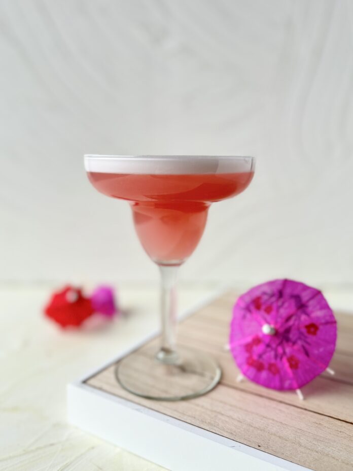 A pink cocktail in a classic margarita glass on a tray with cocktail umbrellas in hot pink and red.
