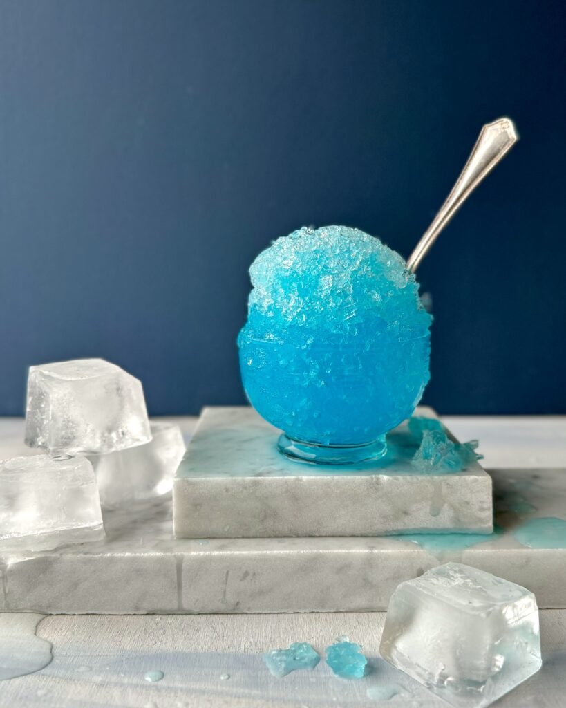 A Zamboni, Snow-Cone Cocktail that's a vibrant blue ball of ice in a clear glass cup with a silver spoon sitting on pieces of white marble with large cubes of ice.