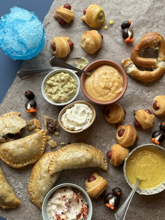 A flat lay photo of variety of handles, pigs in a blanket, soft pretzels with beer cheese, and a blue snow cone cocktail for the best hockey game party menu.