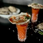 A look in photo of an etched shooter glass with an oyster on the half shell resting on the top of the glass on a black surface, with another oyster shooter in the background along with a dish of oysters.