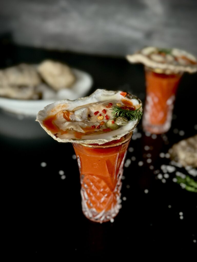 A look in photo of an etched shooter glass with an oyster on the half shell resting on the top of the glass on a black surface, with another oyster shooter in the background along with a dish of oysters.