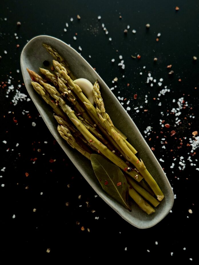 Pickled Asparagus An overhead photo of an oval dish with pickled asparagus spears, a glove of garlic, and a bay leaf, on a black surface with sprinkled salt.