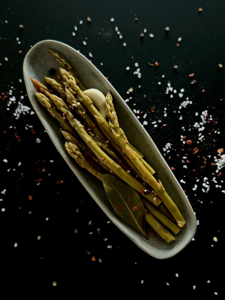 An overhead photo of an oval dish with pickled asparagus spears, a glove of garlic, and a bay leaf, on a black surface with sprinkled salt.