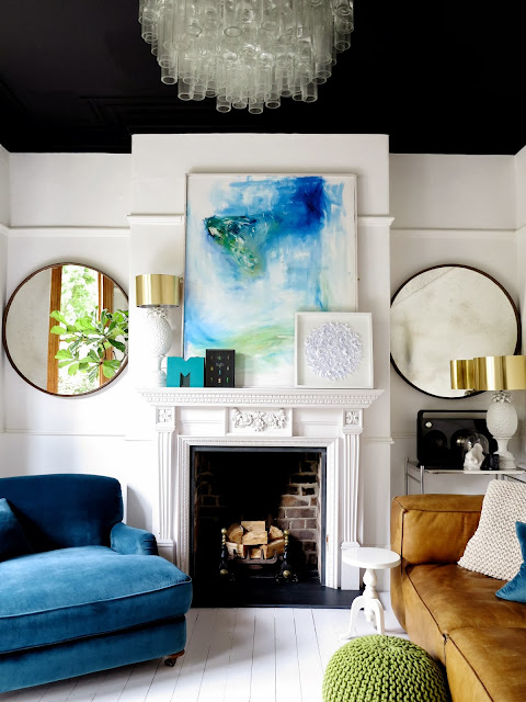 A white walled living room pictures a white fireplace between two big round mirrors with a large painting atop the mantle.
