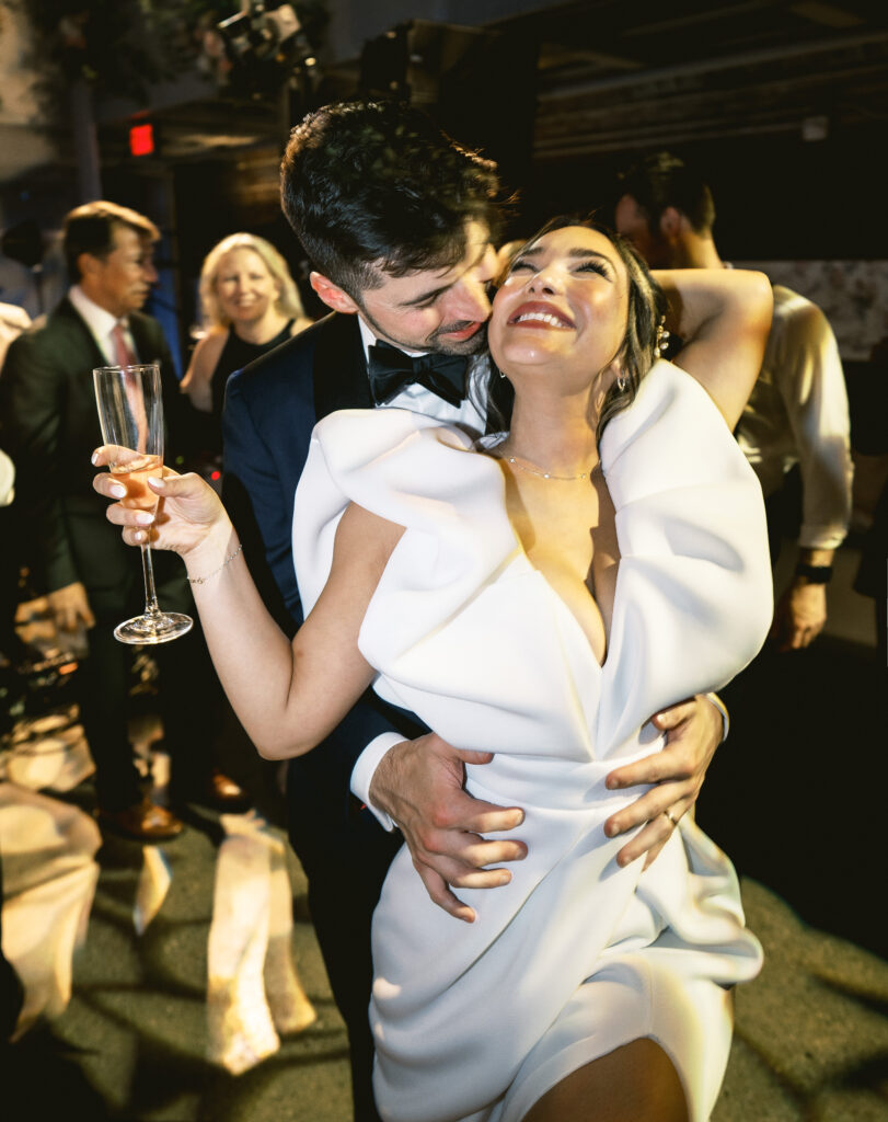 A recently married could dances on the dance floor of their wedding with him in a black suit holding her in her white wedding dress and drink in hand.