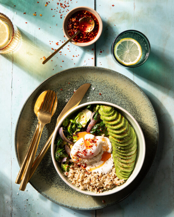 A Quinoa Bowl filled with egg, avocado, kale, and more on a green plate beside a drink, chili oil, and a spoon and knife.