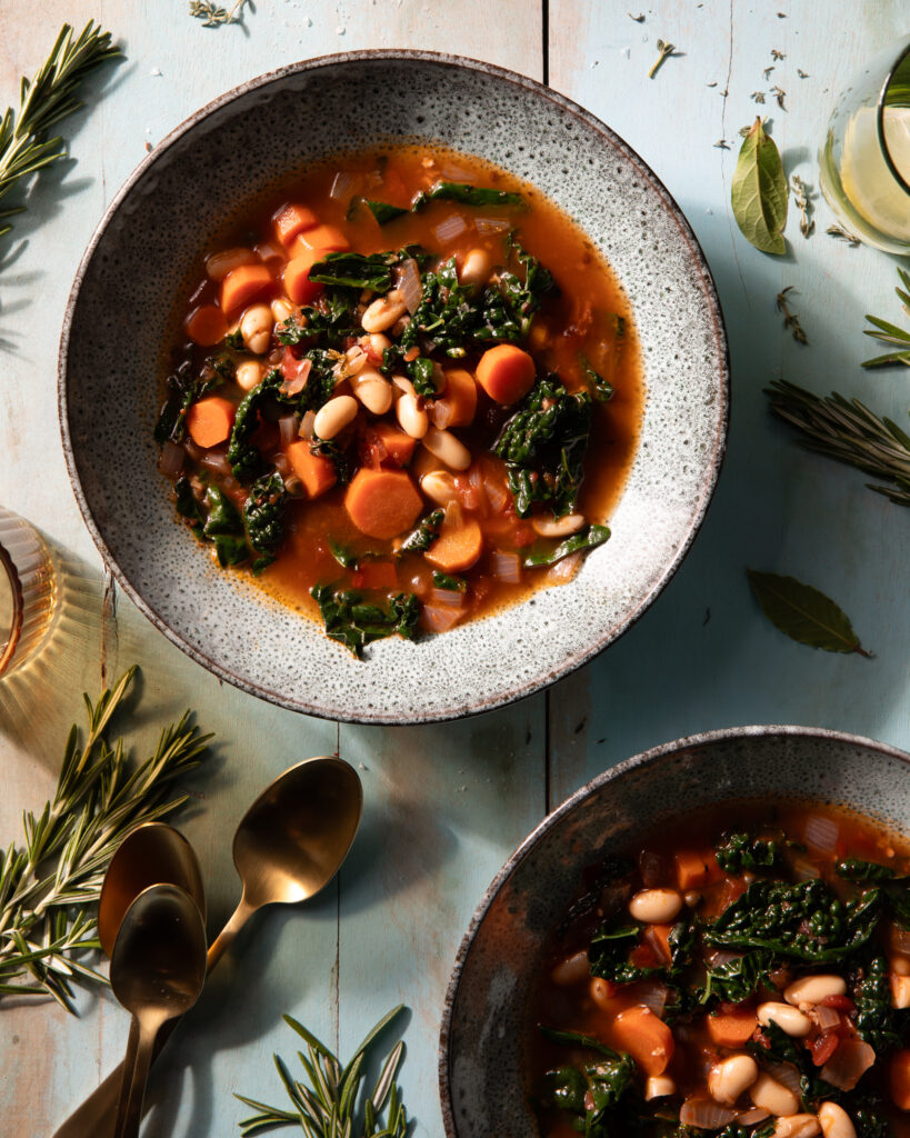 A bowl full of a tomato based Mediterranean Stew with carrots, kale, and beans throughout on top of a wooden table beside a golden spoon.
