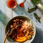 A bowl of sausage, apple sticks, butternut squash, and walnuts with a spoon in it, all laid against a blueish wood table.
