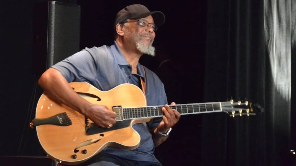 A man in a blue shirt holds a guitar on his lap sporting a grey beard, glasses, and black cap.
