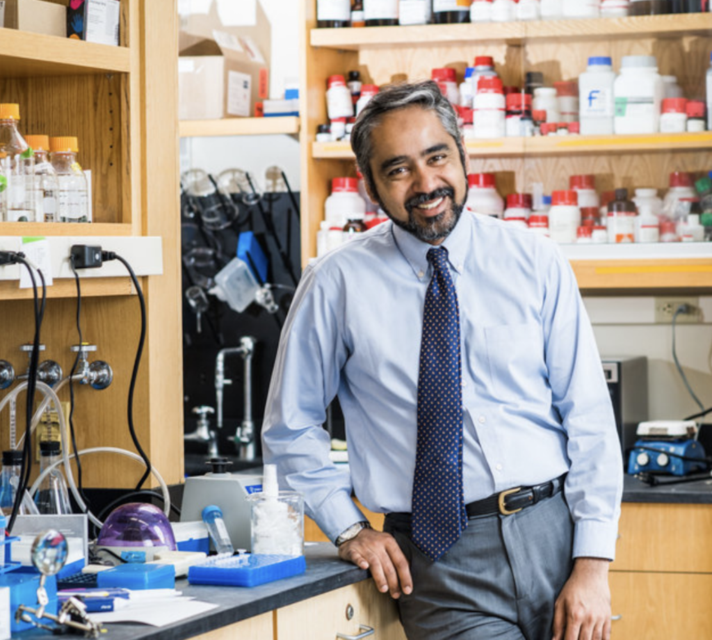 Muhammad Zaman stands beside a lab table filled with supplies in a blue shirt and tie.