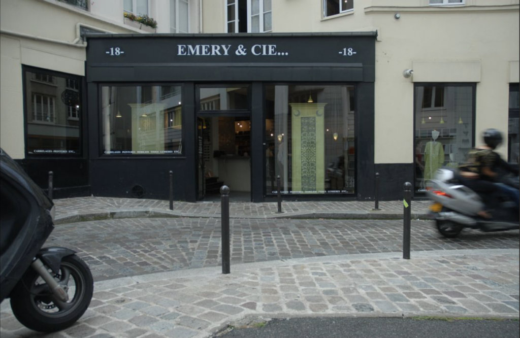 A black storefront for Emery et Cie sits on the stone streets of Paris as mopeds ride by.