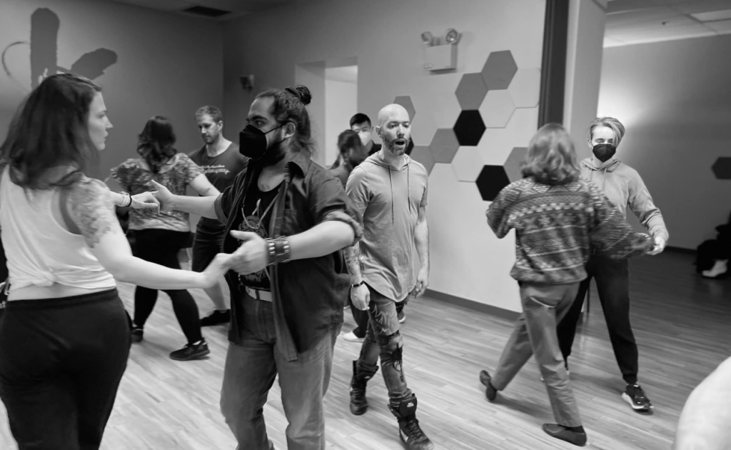 Couples dance around a bald man instructor who walks down the center of the room as the couples dance around him. Photo is in black and white.
