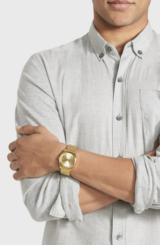 A man in a white shirt has his arms crossed against his body, sporting a gold Nixon watch on his wrist.