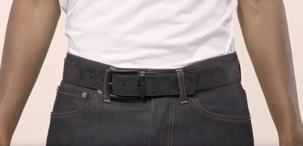 A man in a white shirt and jeans sports a reversible black COACH belt on his waist.
