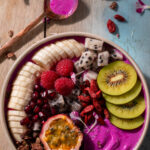 An overhead photo of a colorful tropical smoothie bowl with banana, kiwi, passion fruit, berries, and granola over a vibrant purple yogurt blend in a tan bowl with a gold spoon on a tan and light blue wood surface.