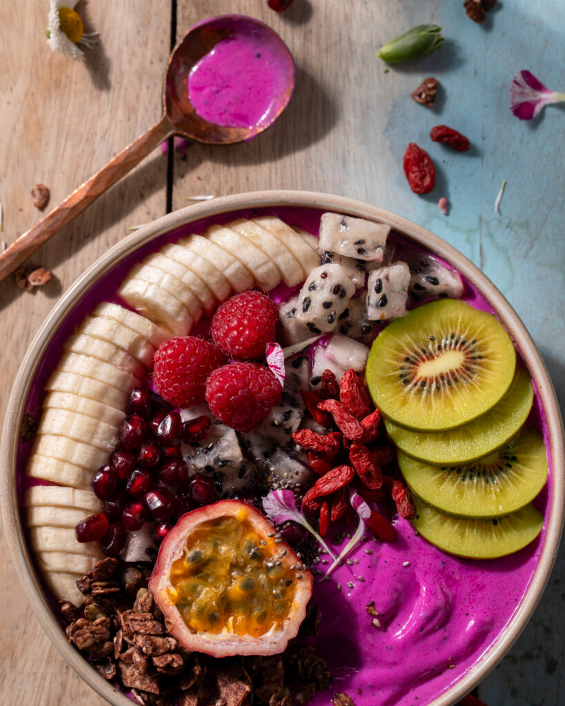 An overhead photo of a colorful tropical smoothie bowl with banana, kiwi, passion fruit, berries, and granola over a vibrant purple yogurt blend in a tan bowl with a gold spoon on a tan and light blue wood surface.