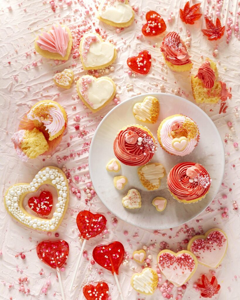A variety of iced cookies, cupcakes and candies all in white, pink and red for Valentine's Day, on a textured white surface with white sprinkles.