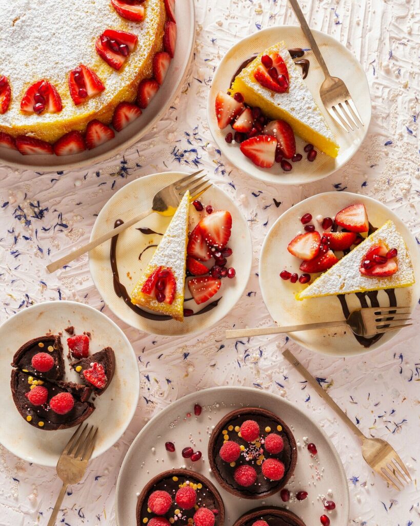 An overhead photo of a Lemon Saffron Tort in the upper left corner, three cream colored plates with slices of the tort with berries, and chocolate tarts at the bottom of the photo.