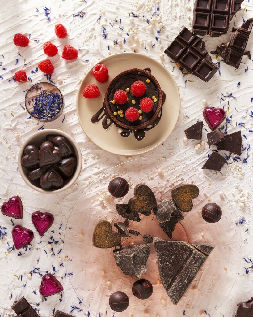 An overhead photograph of chocolate tarts and individual chocolates on a textured white surface with white sprinkles and lavender.