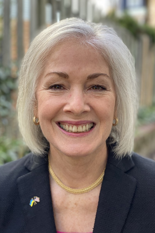 A woman with grey hair wearing a black suit top and gold necklace outside smiles for the camera.