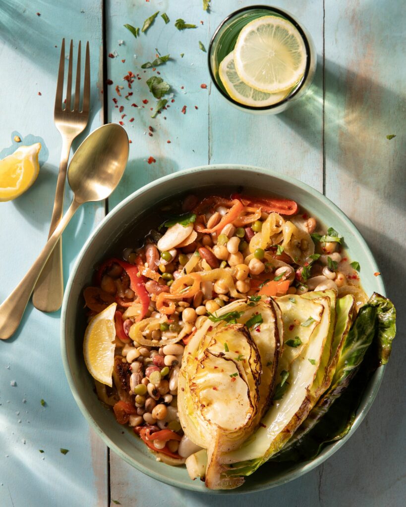 A blue bowl holdsbraised cabbage and beans topped with a lemon slice and accompanied by a fork, spoon, and glass of lemon water.