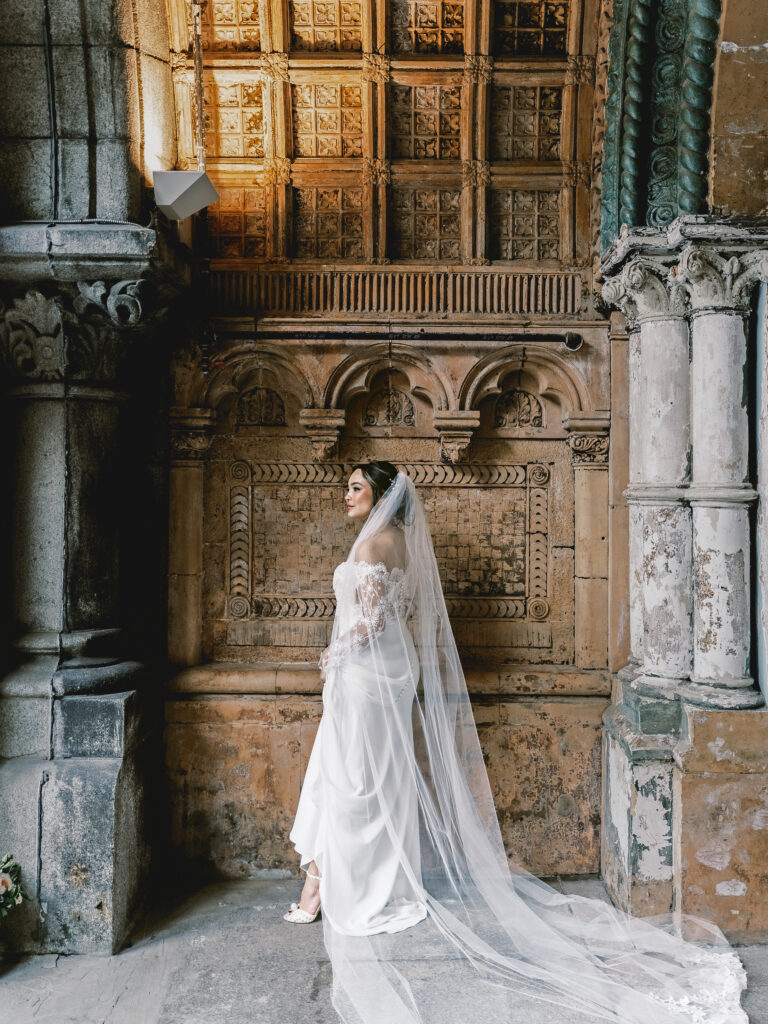 A bride stands in front of an industrial style wall filled with columns in her white wedding dress.