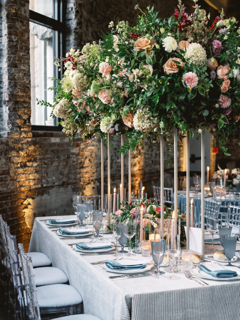 A weddings table set up showcases white table cloths, tall skinny white candles, lots of bouquet flowers, and white table settings.