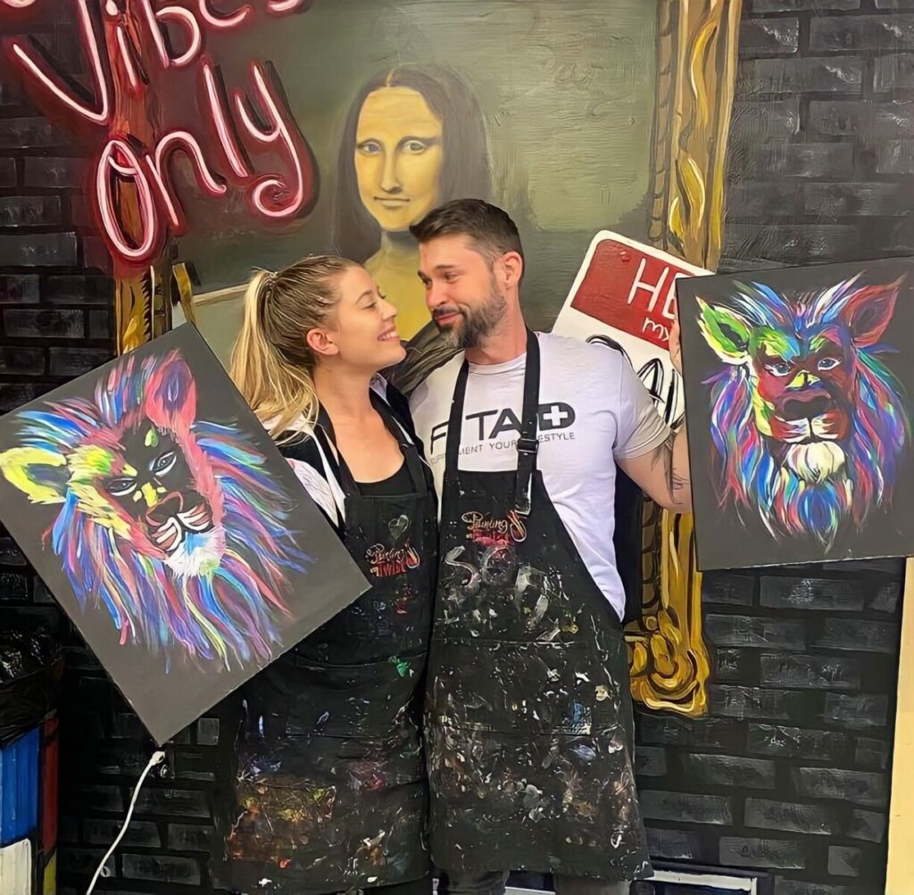 A couple looks at each other with painting aprons on while holding their own painting of a neon lion.