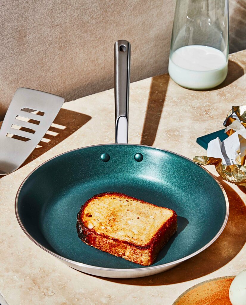 A green pan shwocasing stylish cookware sits on a marble counter with a grilled cheese in the pan and a spatula to the left.